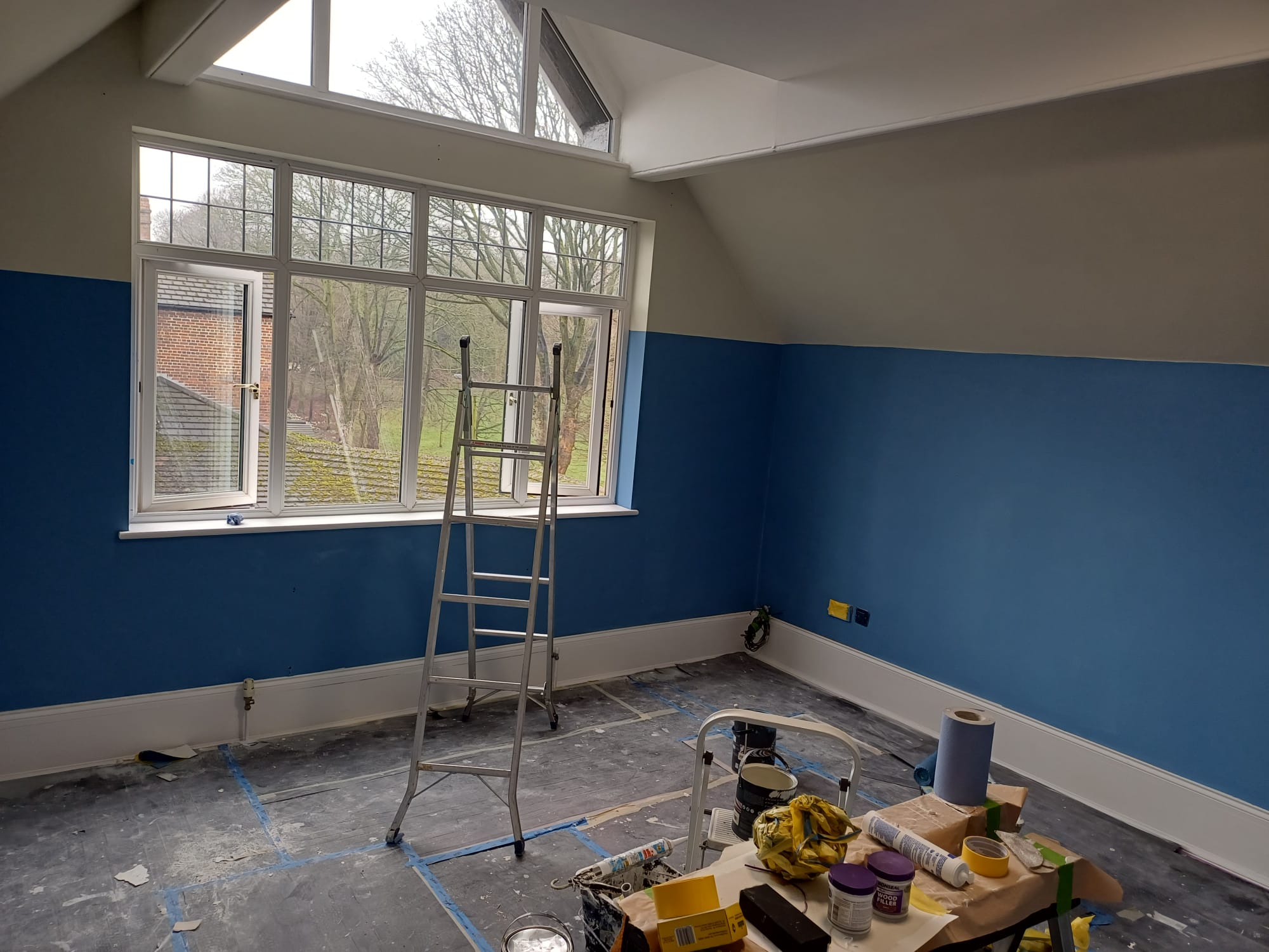 Interior room mid-paint with blue lower walls, cream upper walls, ladder and paint tins