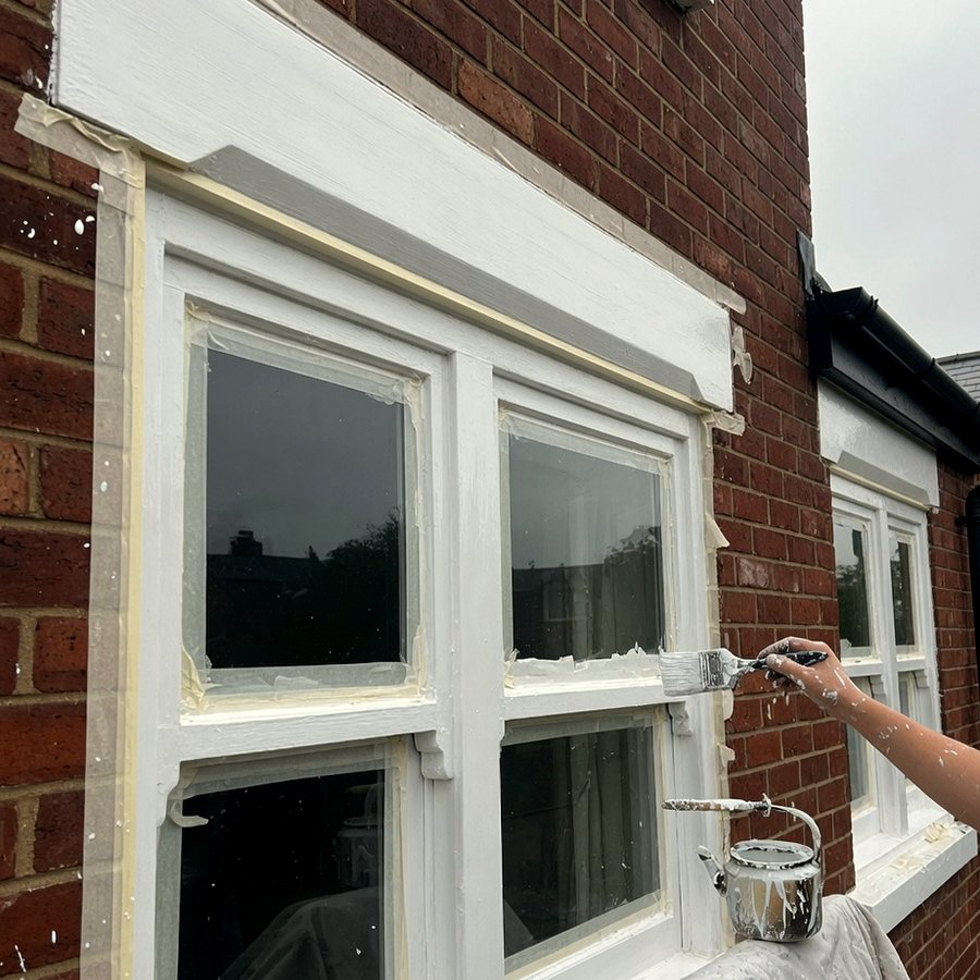 Exterior sash window frames being painted white with a brush and masking tape