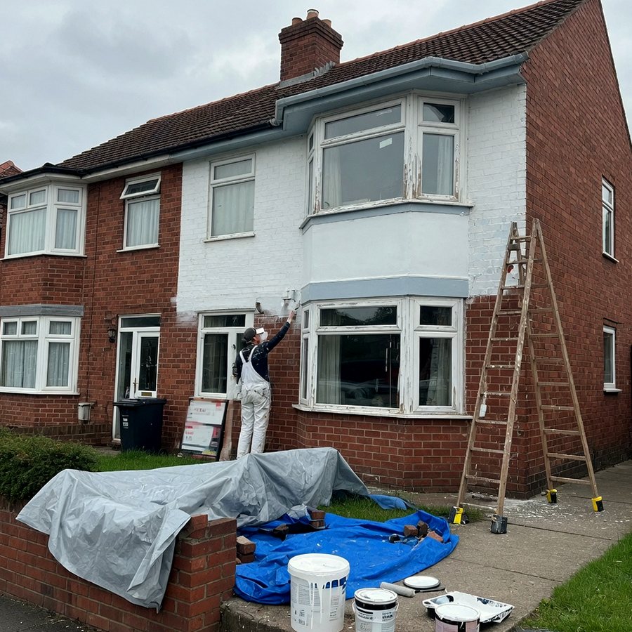 Painter coating the white facade of a red-brick semi, ladder and tarpaulins on the drive